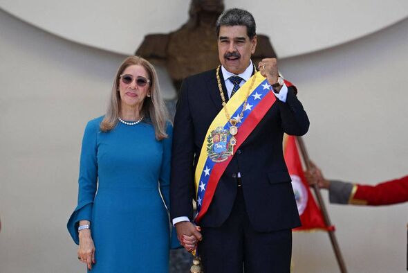 An individual in formal attire, adorned with a sash, stands beside a person in a blue dress, both of them facing forward in a fo