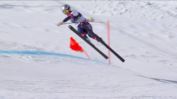 An individual, dressed in a helmet and ski attire, is mid-air on a ski jump, poised above a snowy slope. Nearby, there are sever