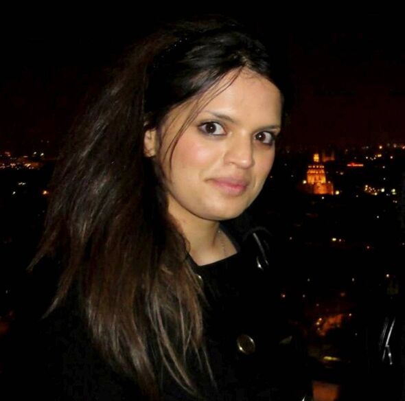 A portrait of an individual with long hair, standing against a dimly lit backdrop featuring illuminated city lights in the dista