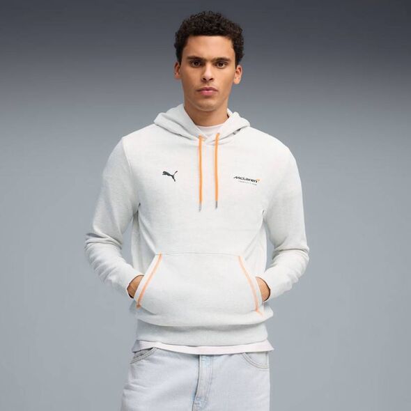 An individual is posing for a photograph while wearing a white hoodie with a logo and a zipper, standing against a neutral backg