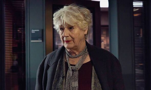 An elderly woman with short, white hair is standing and looking to the left, with a slight frown on her face. She is wearing a d