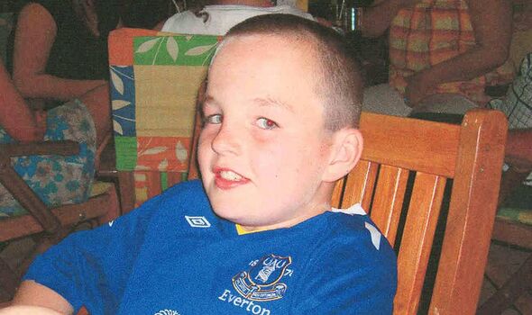 A young boy with a short haircut is seated on a wooden chair, smiling and looking directly at the camera. He is wearing a blue s