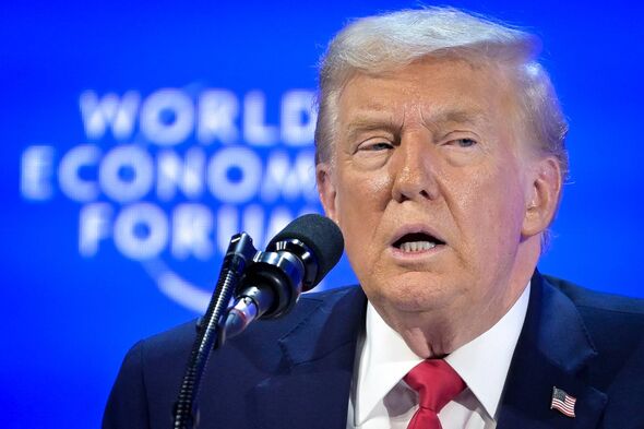 An individual, dressed in formal attire and standing at a podium, delivers a speech in front of a microphone. The backdrop featu