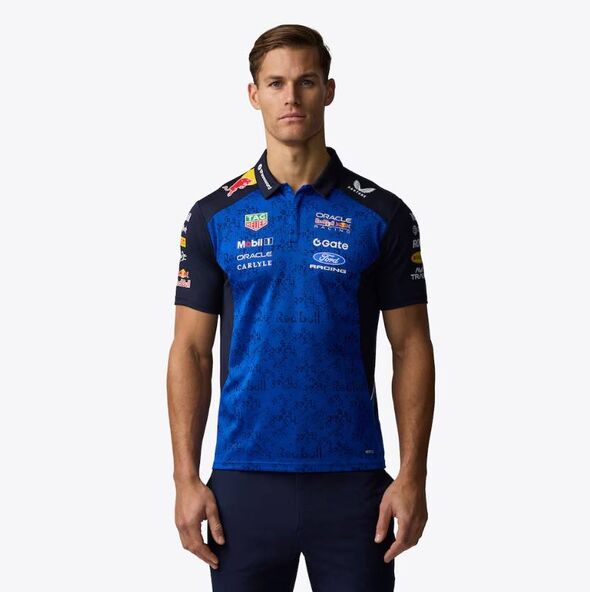 An individual dressed in a blue and black athletic shirt with several logos and sponsor texts is standing and posing for a photo