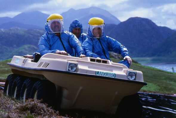 Three individuals dressed in blue protective suits and yellow helmets are riding on a large amphibious vehicle. The vehicle is s