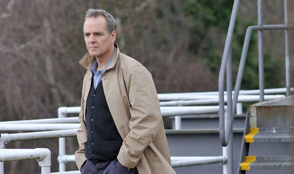 An adult male individual dressed in a black inner garment and a beige overcoat stands on a metallic platform, with his hands in