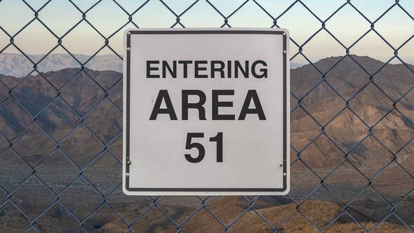 A rectangular sign with white background and black text is hung on a chain-link fence. The sign reads