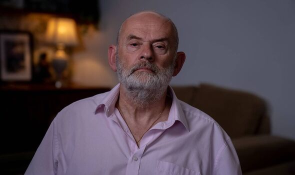 An older gentleman with a white beard and mustache is seated in an indoor setting, wearing a light purple shirt. Behind him, a d