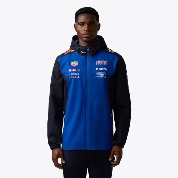 An individual dressed in a blue and black sports jacket with a hood, featuring various sponsor logos and text, poses for a photo