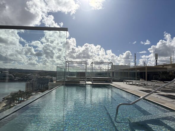 Picture of an outdoor rooftop pool, with the sun shining above in a mostly clear, blue sky