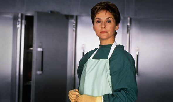 An individual wearing a green jacket and a white apron stands in front of large, metallic refrigerators, their expression contem