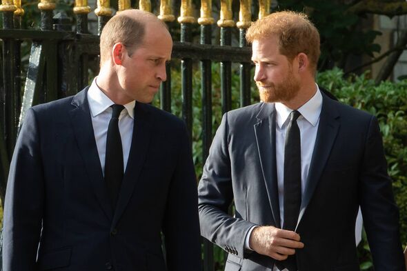 Two individuals dressed in formal attire are engaged in a conversation while standing in front of an ornate, black metal gate.