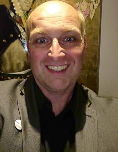 A close-up image of a man with short, light-colored hair, dressed in a dark-colored collared shirt, smiling and appearing to be