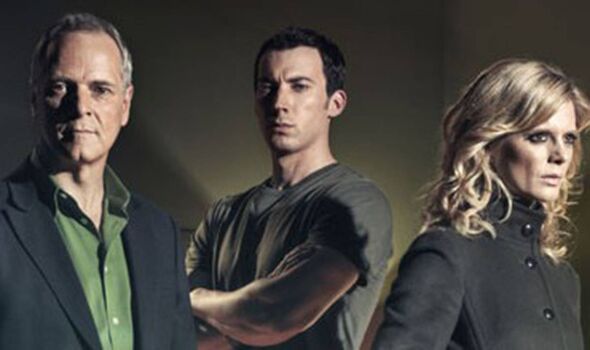 Three individuals are standing together against a plain background. The person on the left is dressed formally in a black suit a