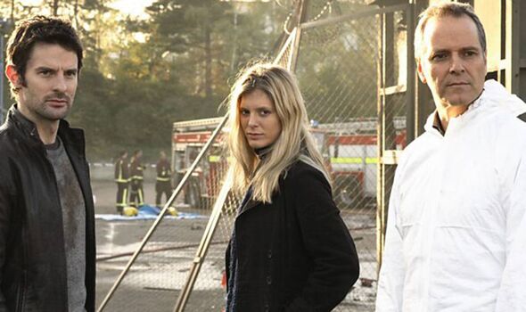 Three individuals are standing in an outdoor setting, with one individual prominently positioned in the center. The background f