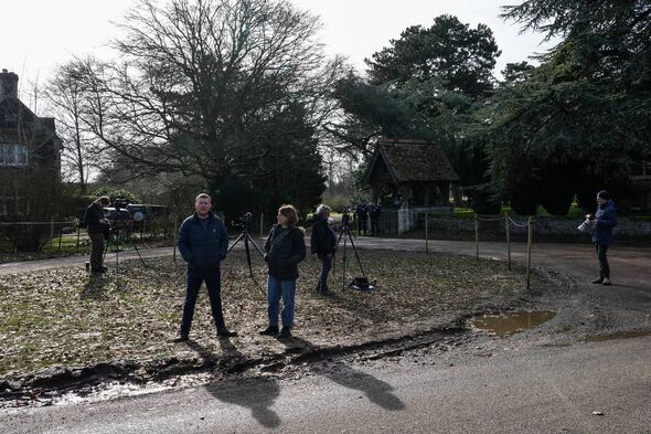 A group of individuals is standing on the side of a road, surrounded by greenery and trees, with a building visible in the backg