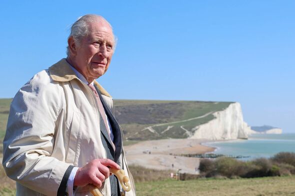 An older gentleman dressed in a light-colored overcoat and a tie stands on a grassy hill under a clear blue sky. Behind him, the