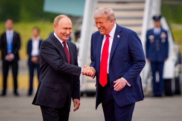 Two men in formal suits, one wearing a blue suit and the other a black suit, are engaged in a handshake while standing on an air