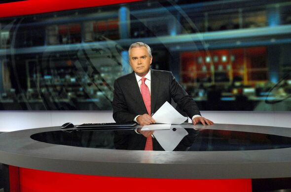 An individual dressed in formal attire, comprising a suit and tie, is seated at a news desk. He holds a piece of paper and appea
