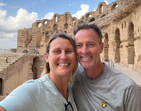 An adult man and woman standing together in front of an ancient ruin, both smiling for a photograph.