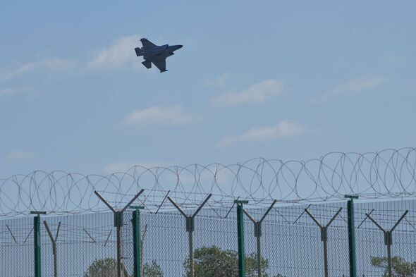 An aerial image captures a jet aircraft soaring above a security fence. The aircraft, positioned against a backdrop of a clear b