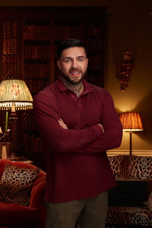 An individual is seated in an interior room, positioned next to a lamp and a decorative wall feature, with a background that inc