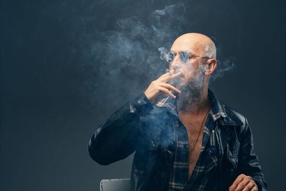 A bald man smoking a cigarette, filling the dimly lit room with smoke