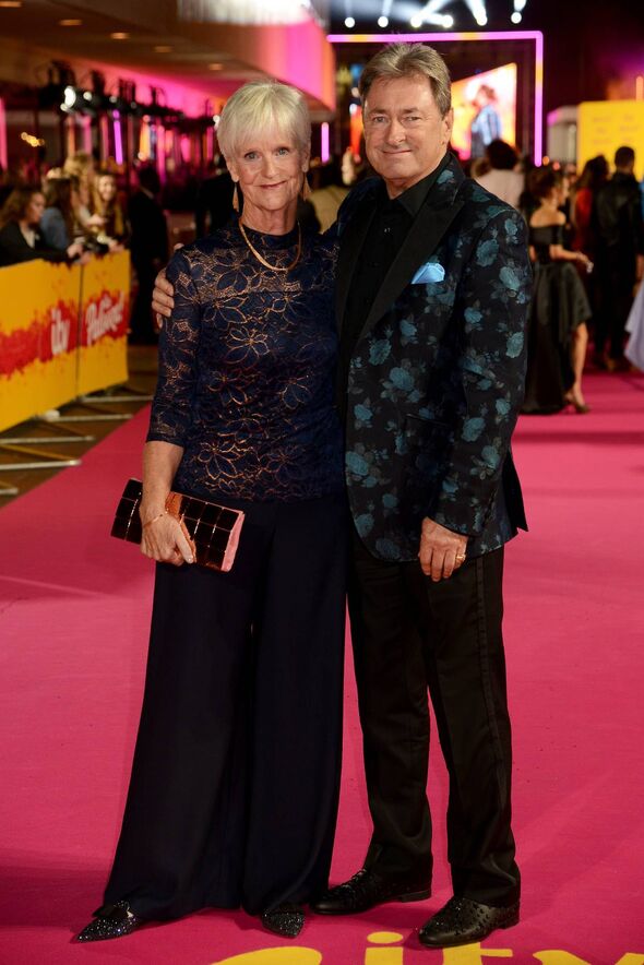 An older woman and a man are standing next to each other on a red carpet, surrounded by a crowd of people and vibrant event deco
