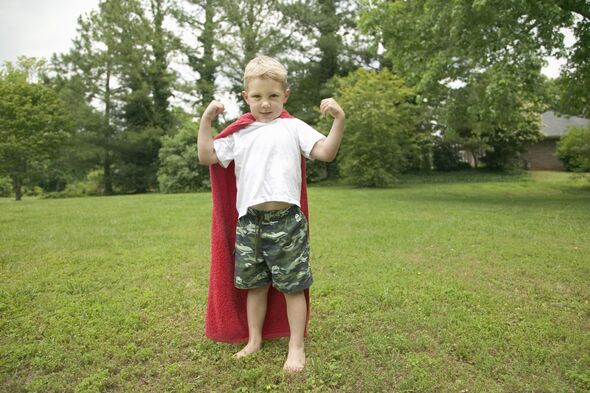 Boy (2-4) standing outdoors on lawn, wearing cape, showing muscles