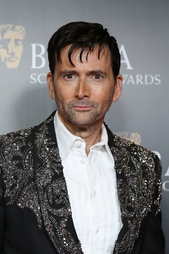 An individual, dressed in a formal suit, standing against a backdrop with the partial text