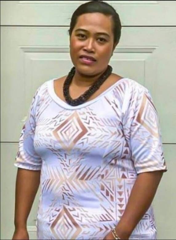 An adult female individual dressed in a white shirt adorned with floral patterns and a black necklace stands in front of a plain
