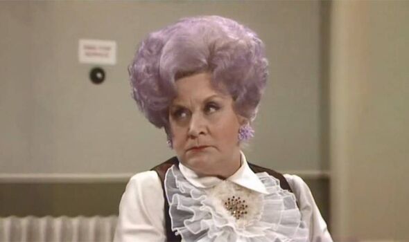 An individual with an eccentric appearance, donning a white blouse with ruffles and a contrasting black vest, stands in an indoo