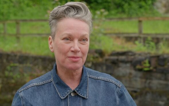An adult individual with short gray hair standing in front of a rustic stone wall and a natural green landscape in the backgroun