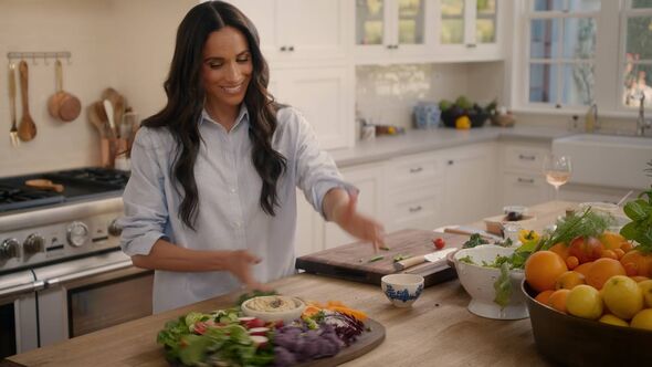 An individual with long hair, dressed in a light-colored shirt, is engaged in food preparation within a kitchen environment. Var