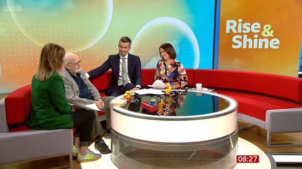 A formal gathering takes place in a studio setting, featuring a group of individuals seated on a red couch. They are engaged in