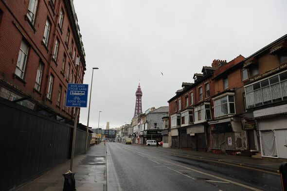 An empty urban street with multi-story buildings on either side, featuring a blue sign and a distant tower, under an overcast sk
