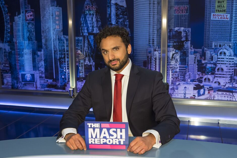 Nish Kumar sits at a newsdesk holding a card with The Mash Report branding on it
