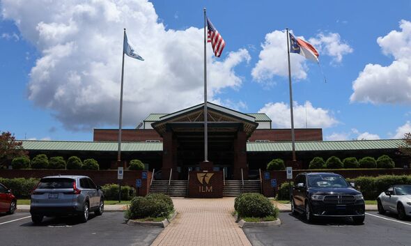 An exterior view of a building with flags atop flagpoles, cars parked in the driveway, and a walkway leading to the entrance.