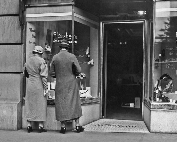 A black-and-white photograph depicts two individuals standing in front of a store. They appear to be examining the shop's window