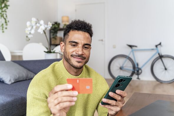 Young adult man smiling, holding credit card and smartphone for online shopping and secure mobile payment