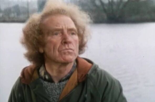 An individual with curly, light-colored hair wearing a green jacket and a brown strap around their neck is standing against the