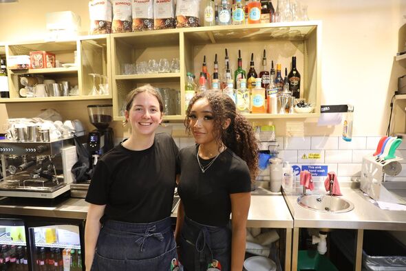 Two individuals are standing in front of a counter in what appears to be a café setting, with various items and appliances visib