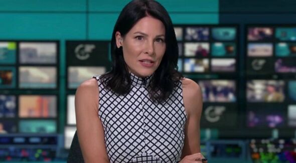 An individual, possibly a journalist or presenter, stands before a wall adorned with multiple screens displaying various images