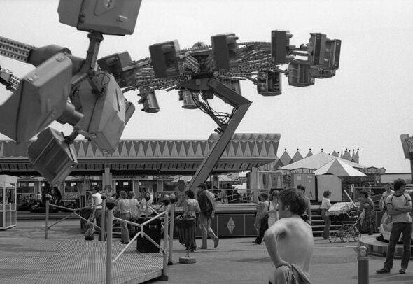 A black-and-white photograph depicts an amusement park with a large mechanical ride featuring multiple rotating arms and seats.