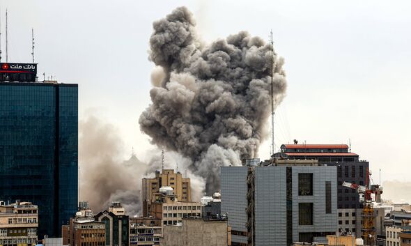 An aerial view depicts a large plume of smoke and dust rising from an urban area, with several buildings visible in the foregrou