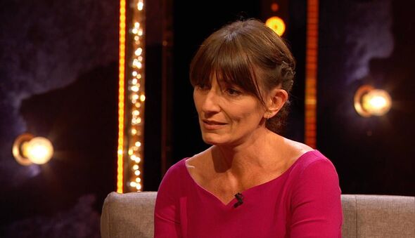 An individual with short hair and wearing a pink top is seated on a couch, possibly engaged in an interview or discussion.