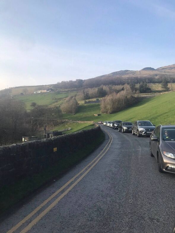 A scenic rural road flanked by verdant hills and trees, with a line of vehicles moving slowly along the route.