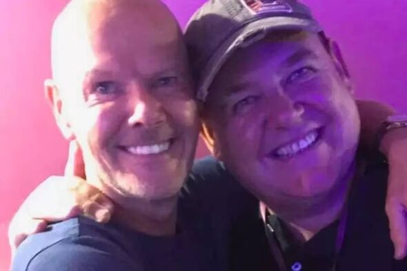 Two individuals, one bald and the other sporting a cap, are smiling and posing closely together against a vibrant purple backdro