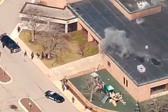 An aerial view depicts a building emitting smoke, with several vehicles parked nearby and individuals present in the vicinity.