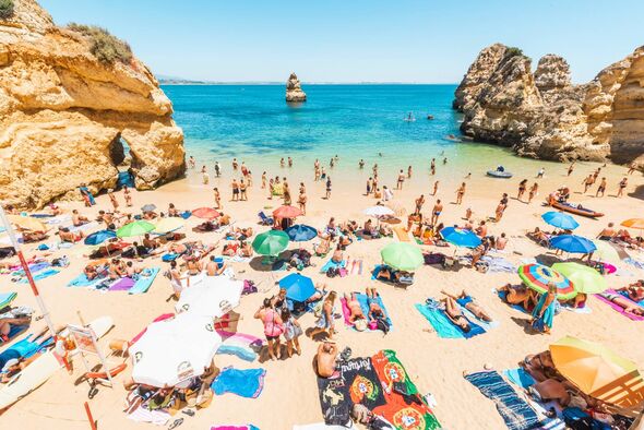 An aerial view of a crowded beach with numerous individuals and beach umbrellas. The beach is bordered by a clear blue ocean and